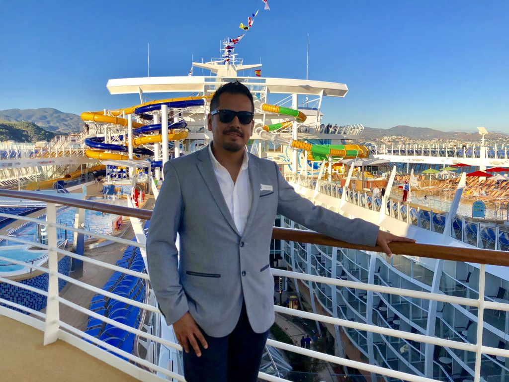 Carlos Perez, Cruise Recruitment Manager, during his time working onboard a yacht. Stood on the deck balcony in front of water park. 